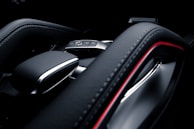 A close-up image of a car's interior focusing on the gear shift area. The design is modern and sleek, featuring a leather finish and metallic accents. A small key fob with buttons is visible, indicating functions such as lock and unlock.