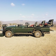 A green safari vehicle with open seating and sturdy build, parked on dry, dusty terrain with a scenic backdrop of distant hills under a clear blue sky.