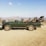 A green safari vehicle with open seating and sturdy build, parked on dry, dusty terrain with a scenic backdrop of distant hills under a clear blue sky.