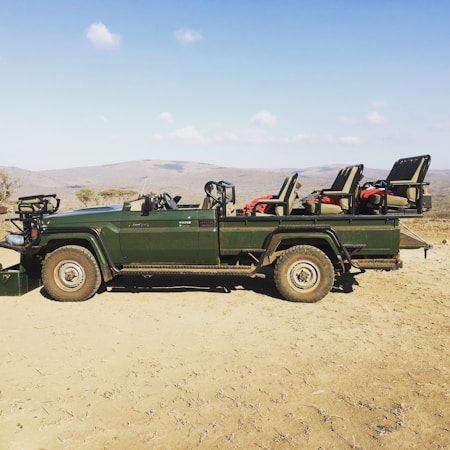 A green safari vehicle with open seating and sturdy build, parked on dry, dusty terrain with a scenic backdrop of distant hills under a clear blue sky.