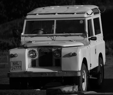 A vintage off-road vehicle, likely a Land Rover, is parked on a paved surface. The vehicle is an older model characterized by its boxy shape, large round headlights, and utility-style features. It has a classic grille and a roof with added structures. The image is black and white, emphasizing the vehicle's details and textures.