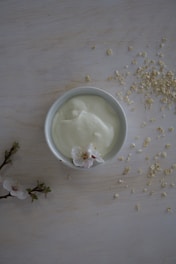 A small bowl filled with creamy yogurt is placed on a pale wooden surface. Scattered around the bowl are flakes of oats, adding a rustic touch. A delicate flower with soft white petals and a pink center is positioned on the side of the bowl. In the lower left corner, a branch with a few more flowers and budding leaves complements the scene.