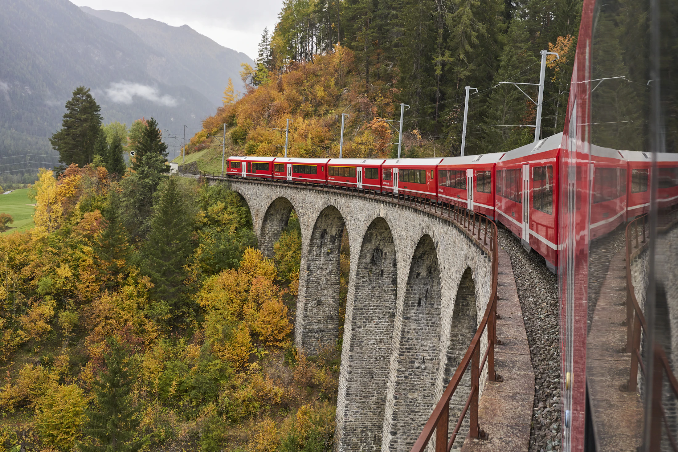 A red Rhaetian Railway train crossing a stone viaduct on the Albula line in autumn