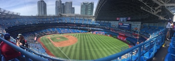 A large baseball stadium with a partially open roof, green grass field, and red clay infield. Buildings are visible in the background, and spectators are seated in blue seats. A few banners and flags are displayed, including a scoreboard to the right.