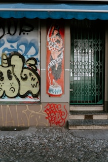 A storefront displays an illustrated panel of a woman in a polka-dot dress and top hat, titled 'Zirkus'. The adjacent wall and window feature graffiti art with abstract patterns in blue, yellow, and black. The entrance has a closed metal gate with steps leading up to it, and red graffiti is prominently visible at the bottom.
