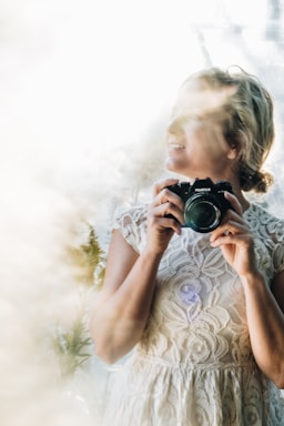 A person in a lace dress holds a camera, standing in front of a bright, softly focused background. The light creates a dreamy atmosphere with a mix of natural elements and soft shadows.