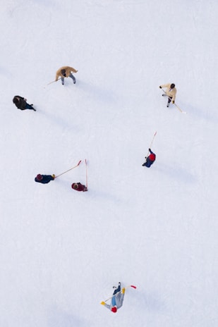 An aerial view of several people on an ice rink. They are playing with sticks, likely engaged in a friendly ice hockey game. The ice is smooth and white, providing a contrasting background to the colorful clothing and equipment.
