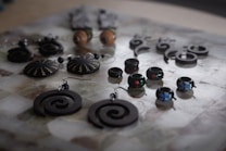 An assortment of handcrafted jewelry displayed on a surface, featuring a variety of earrings and rings with intricate designs. The pieces include spiral-shaped earrings, round earrings with zigzag patterns, and small rings adorned with colorful designs. The jewelry is mostly made of dark materials, creating a striking contrast with the lighter background.