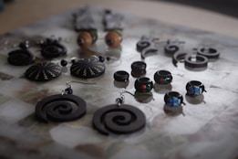 An assortment of handcrafted jewelry displayed on a surface, featuring a variety of earrings and rings with intricate designs. The pieces include spiral-shaped earrings, round earrings with zigzag patterns, and small rings adorned with colorful designs. The jewelry is mostly made of dark materials, creating a striking contrast with the lighter background.