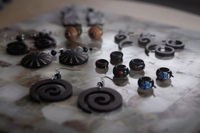 An assortment of handcrafted jewelry displayed on a surface, featuring a variety of earrings and rings with intricate designs. The pieces include spiral-shaped earrings, round earrings with zigzag patterns, and small rings adorned with colorful designs. The jewelry is mostly made of dark materials, creating a striking contrast with the lighter background.
