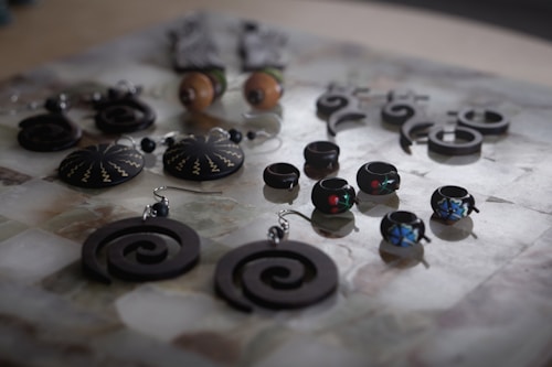 An assortment of handcrafted jewelry displayed on a surface, featuring a variety of earrings and rings with intricate designs. The pieces include spiral-shaped earrings, round earrings with zigzag patterns, and small rings adorned with colorful designs. The jewelry is mostly made of dark materials, creating a striking contrast with the lighter background.