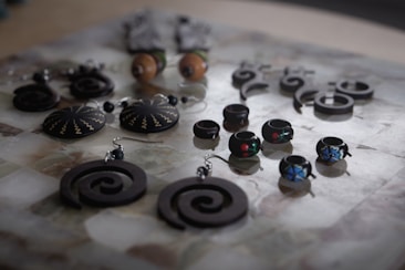 An assortment of handcrafted jewelry displayed on a surface, featuring a variety of earrings and rings with intricate designs. The pieces include spiral-shaped earrings, round earrings with zigzag patterns, and small rings adorned with colorful designs. The jewelry is mostly made of dark materials, creating a striking contrast with the lighter background.