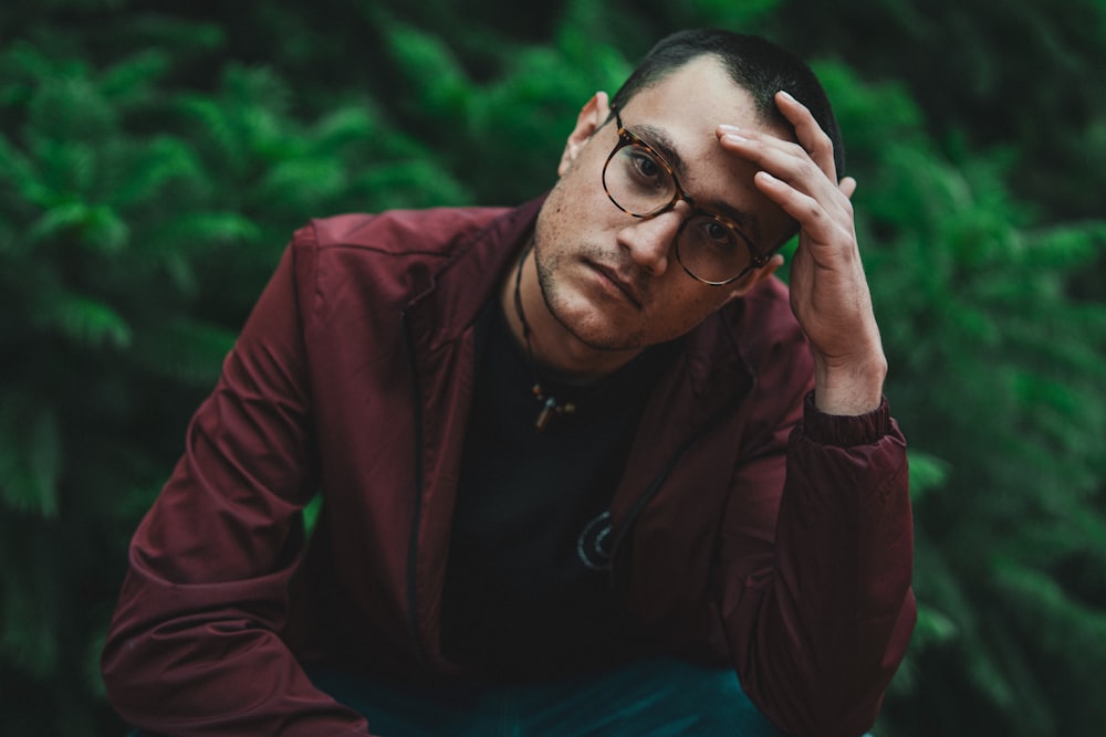 man wearing red collared button-up long-sleeved shirt and eyeglasses sitting while touching his forehead