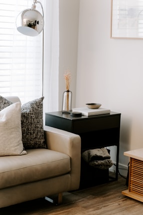 A cozy living room corner featuring a beige couch with a mix of textured pillows. Next to the couch, a sleek black side table holds a decorative vase with dried flowers and a few books. A modern metallic lamp hangs overhead, casting a gentle glow on the scene, while light streams in through white blinds on the window.