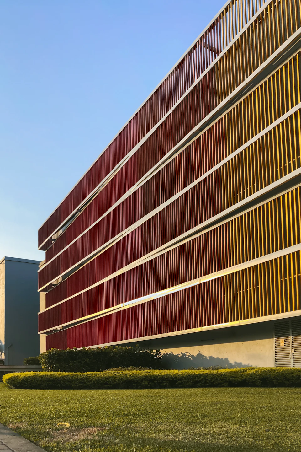 The image shows a modern, multi-story building with a unique facade composed of red and yellow panels arranged in a zigzag pattern. The building is situated on a street corner, surrounded by lush green grass that contrasts with its vibrant colors. In the foreground, there are trees providing additional greenery to the scene.