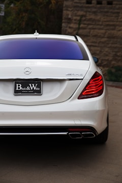 A rear view of a sleek white luxury car with illuminated red taillights. The car includes an AMG badge, a prominent exhaust system, and a black license plate that reads 'Black & White'. The background features a stone wall and some greenery.