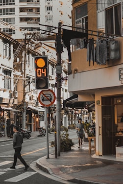 A city street scene with a pedestrian crossing the road. An orange countdown traffic signal is visible, along with a no-parking sign. Buildings with hanging laundry and a mix of old and modern architecture create a dynamic urban setting. Vegetation lines the sidewalk, and a few people are walking or riding motorcycles.