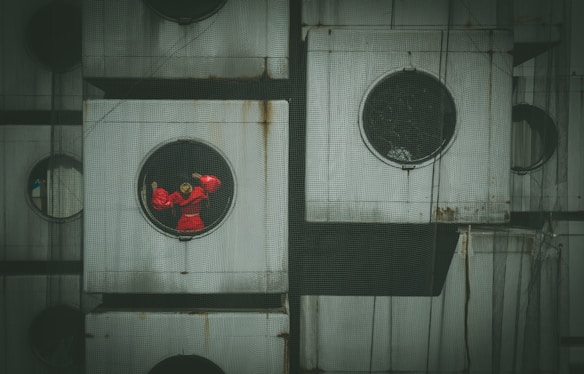 Several square units with circular windows, one of which contains a figure wearing bright red clothing. The architectural style is modular with a grid-like structure. There is a netting covering the entire facade, adding an additional layer of texture.