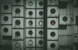 A detailed close-up of a building facade composed of uniformly stacked, cube-like modules with circular windows. Each module is weathered and appears aged, with one window revealing a person in a bright red outfit.