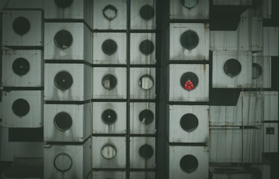 A detailed close-up of a building facade composed of uniformly stacked, cube-like modules with circular windows. Each module is weathered and appears aged, with one window revealing a person in a bright red outfit.