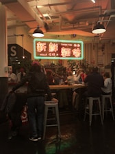 A bustling indoor food court or dining area features a large neon sign with both English and Chinese text hanging above a group of people seated at a communal table. The setting is warmly lit, and there are plants arranged around the space. Various diners are engaged in conversation as they enjoy their meals.