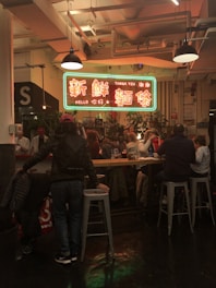 A bustling indoor food court or dining area features a large neon sign with both English and Chinese text hanging above a group of people seated at a communal table. The setting is warmly lit, and there are plants arranged around the space. Various diners are engaged in conversation as they enjoy their meals.