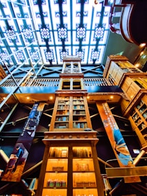 An intricate library interior with a tall wooden bookshelf reaching up towards a stained-glass ceiling. The bookshelf is filled with various books and is surrounded by decorative banners featuring artwork and text. The lighting is warm, providing a cozy atmosphere, while the ceiling's geometric pattern adds a modern touch.
