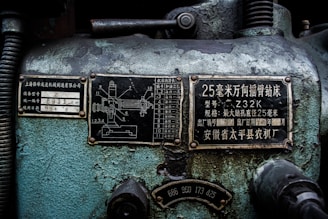 A close-up view of an industrial machine panel with various Chinese characters and numerical labels. The surface displays wear and crackling paint, suggesting age and use. There are two metal plates with technical diagrams and descriptions, along with numbers and letters in white on a black background.