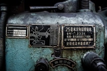 A close-up view of an industrial machine panel with various Chinese characters and numerical labels. The surface displays wear and crackling paint, suggesting age and use. There are two metal plates with technical diagrams and descriptions, along with numbers and letters in white on a black background.