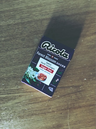 A package of Ricola Forest Blossom Swiss Herb Lozenges is placed on a wooden surface. The packaging features floral imagery and text indicating it is sugar-free.