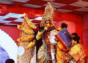 A richly decorated statue of a deity with multiple arms is surrounded by a group of people. The statue is adorned with intricate ornaments and vibrant attire. The people around the statue appear to be performing a ritual or paying their respects, dressed in colorful traditional garments. The setting looks festive with bright red and white canopies overhead.