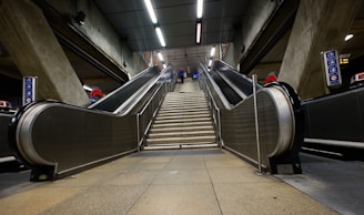 Elevators and a central staircase lead upwards in a modern and industrial underground train station setting. The structural concrete walls flank the escalators, and overhead fluorescent lighting provides illumination. Signs with subway symbols are visible on either side.
