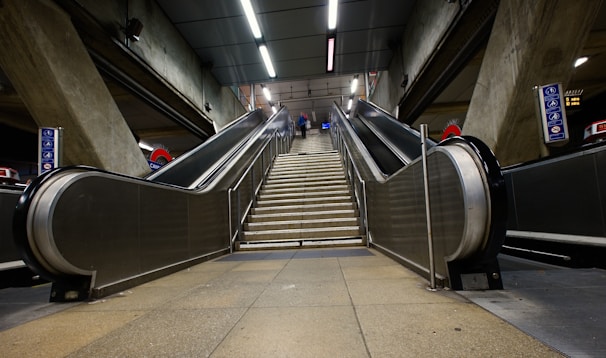 Elevators and a central staircase lead upwards in a modern and industrial underground train station setting. The structural concrete walls flank the escalators, and overhead fluorescent lighting provides illumination. Signs with subway symbols are visible on either side.