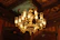 An ornate chandelier with multiple lit candle-like bulbs hangs from a ceiling. The chandelier is elaborately designed with intricate gold detailing and clear glass shades. The ceiling is richly decorated with wood paneling and artistic designs, including floral and foliage patterns. The warm golden glow from the chandelier creates a cozy and elegant atmosphere.