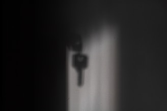 A soft shadow of a key is projected onto a wall, with the shadow varying in darkness and sharpness. The surrounding area is predominantly dark, creating a mysterious or contemplative atmosphere.