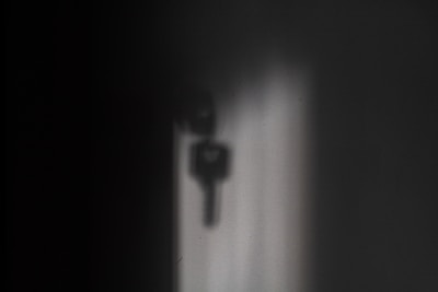 A soft shadow of a key is projected onto a wall, with the shadow varying in darkness and sharpness. The surrounding area is predominantly dark, creating a mysterious or contemplative atmosphere.