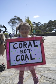 A young girl is smiling while holding a colorful protest sign that reads 'CORAL NOT COAL'. She's outdoors on a sunny day, with trees and a few people visible in the background.