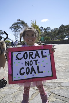 A young girl is smiling while holding a colorful protest sign that reads 'CORAL NOT COAL'. She's outdoors on a sunny day, with trees and a few people visible in the background.