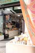 A small shop with a variety of products displayed. Vegetables and fruits are positioned near the entrance, along with some packaged goods. The shop interior is filled with various items, and a person is seen inside near the counter. A banner hangs at the entrance with Japanese writing. The shop has a welcoming atmosphere with natural sunlight illuminating the entrance.