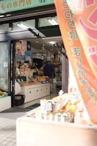 A small shop with a variety of products displayed. Vegetables and fruits are positioned near the entrance, along with some packaged goods. The shop interior is filled with various items, and a person is seen inside near the counter. A banner hangs at the entrance with Japanese writing. The shop has a welcoming atmosphere with natural sunlight illuminating the entrance.