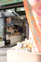 A small shop with a variety of products displayed. Vegetables and fruits are positioned near the entrance, along with some packaged goods. The shop interior is filled with various items, and a person is seen inside near the counter. A banner hangs at the entrance with Japanese writing. The shop has a welcoming atmosphere with natural sunlight illuminating the entrance.