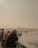 An elderly man leans against a railing, looking out over a body of water. Several other people are nearby, some engaged in fishing. Boats can be seen on the water, and a cityscape with domed buildings is visible in the hazy background.