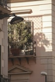 A small balcony with wrought iron railings holds a potted plant with green foliage in front of a set of wooden shutters. Sunlight casts intricate shadows on the light-colored building facade, with an overhead street lamp in the foreground.