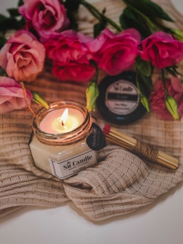 A lit candle in a glass jar labeled 'Soy Candle' rests on a textured beige cloth. Surrounding the candle are vibrant pink roses with lush green leaves. In the background, a jar of matches is visible, adding to the cozy ambiance.