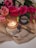 A lit candle in a glass jar labeled 'Soy Candle' rests on a textured beige cloth. Surrounding the candle are vibrant pink roses with lush green leaves. In the background, a jar of matches is visible, adding to the cozy ambiance.