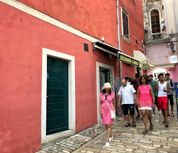 A group of people walks along a narrow street with a pink wall on their left. There is a sign for a currency exchange shop above a door. The street is paved with stone tiles, and people are dressed in casual summer clothing.