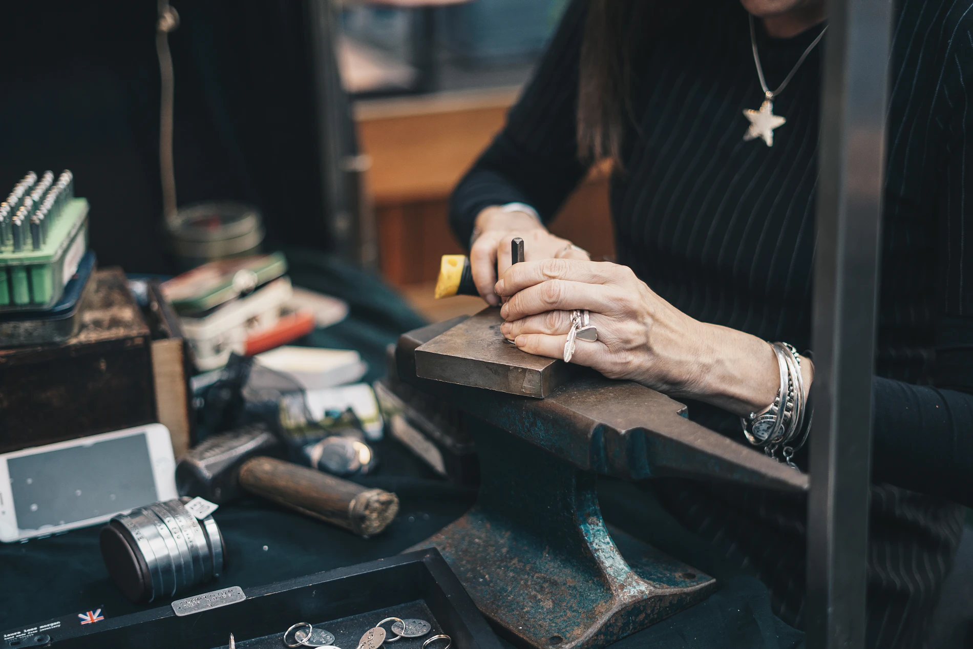A silversmith's hands hold a punch steady against a steel block on the anvil, rings on her fingers, hammers and silver blanks laid out around her.