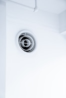 A circular ventilation duct is embedded in a white wall, featuring a series of concentric rings inside the vent. The surrounding area is clean and minimalistic with an overall bright appearance.