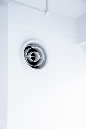 A circular ventilation duct is embedded in a white wall, featuring a series of concentric rings inside the vent. The surrounding area is clean and minimalistic with an overall bright appearance.
