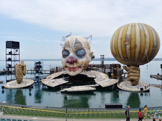A large outdoor stage setup on a body of water features an oversized clown face. On either side of the stage, there are giant sculptural hands, one holding a balloon. The stage is surrounded by water and there is seating available in the foreground.